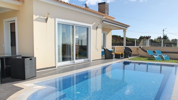 Una piscina con solárium de baldosas, un porche cubierto con sillones de descanso y una entrada con puerta corrediza de vidrio.