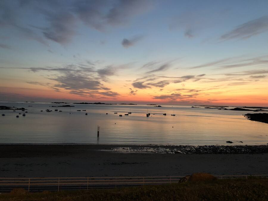 A coastal landscape with boats on calm waters, a sandy beach, and a metal fence in the foreground.