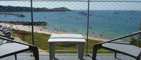 A balcony with a table and chairs overlooking a beach with boats and people.
