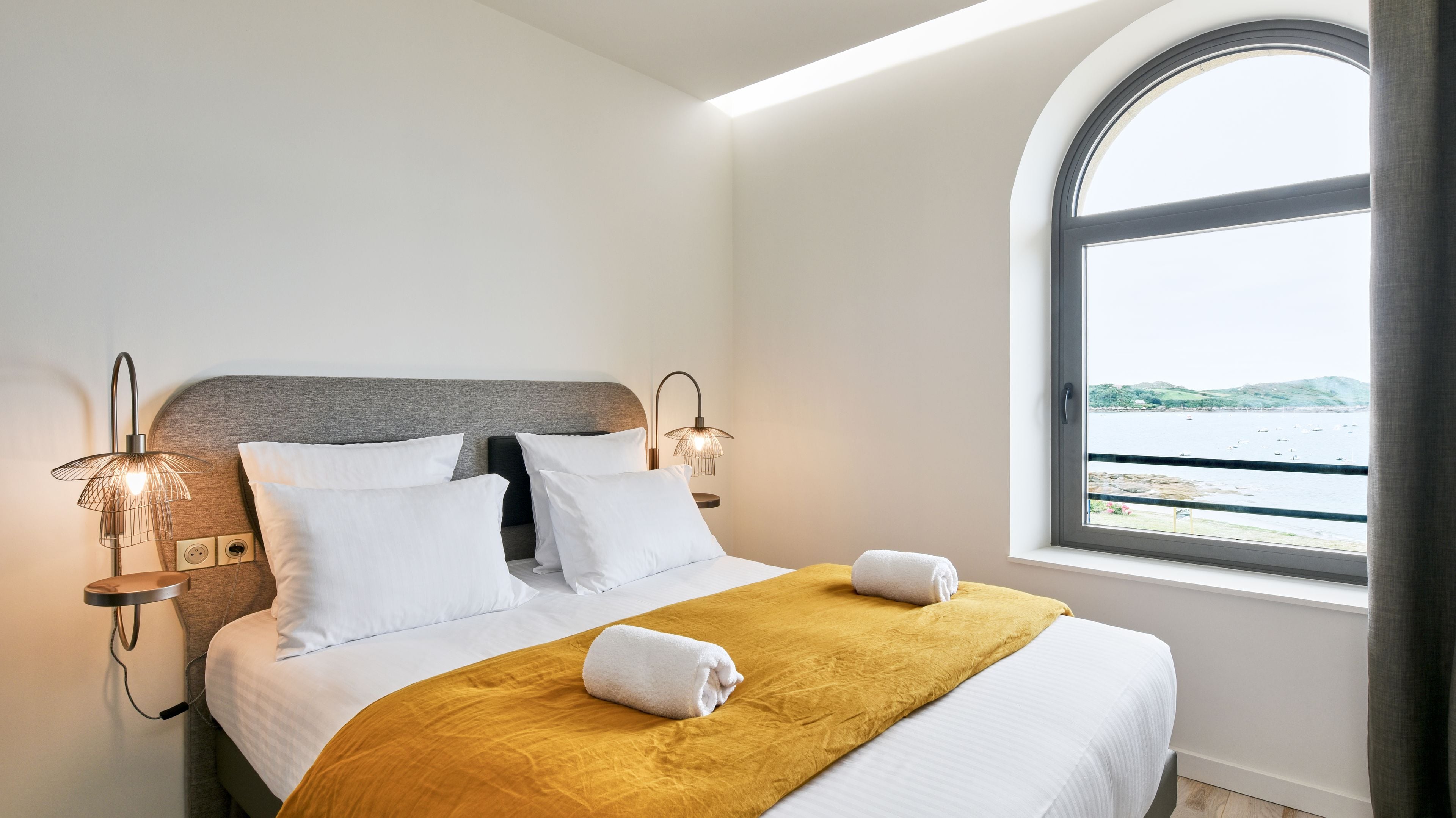 Habitación de hotel con cama, dos lámparas de pared, una ventana con vista al mar y una manta amarilla.