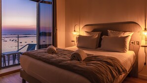 Habitación de hotel con una cama grande, dos lámparas de noche y vista a la playa por una ventana abierta.