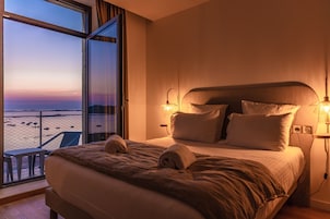 Habitación de hotel con una cama grande, dos lámparas de noche y vista a la playa por una ventana abierta.