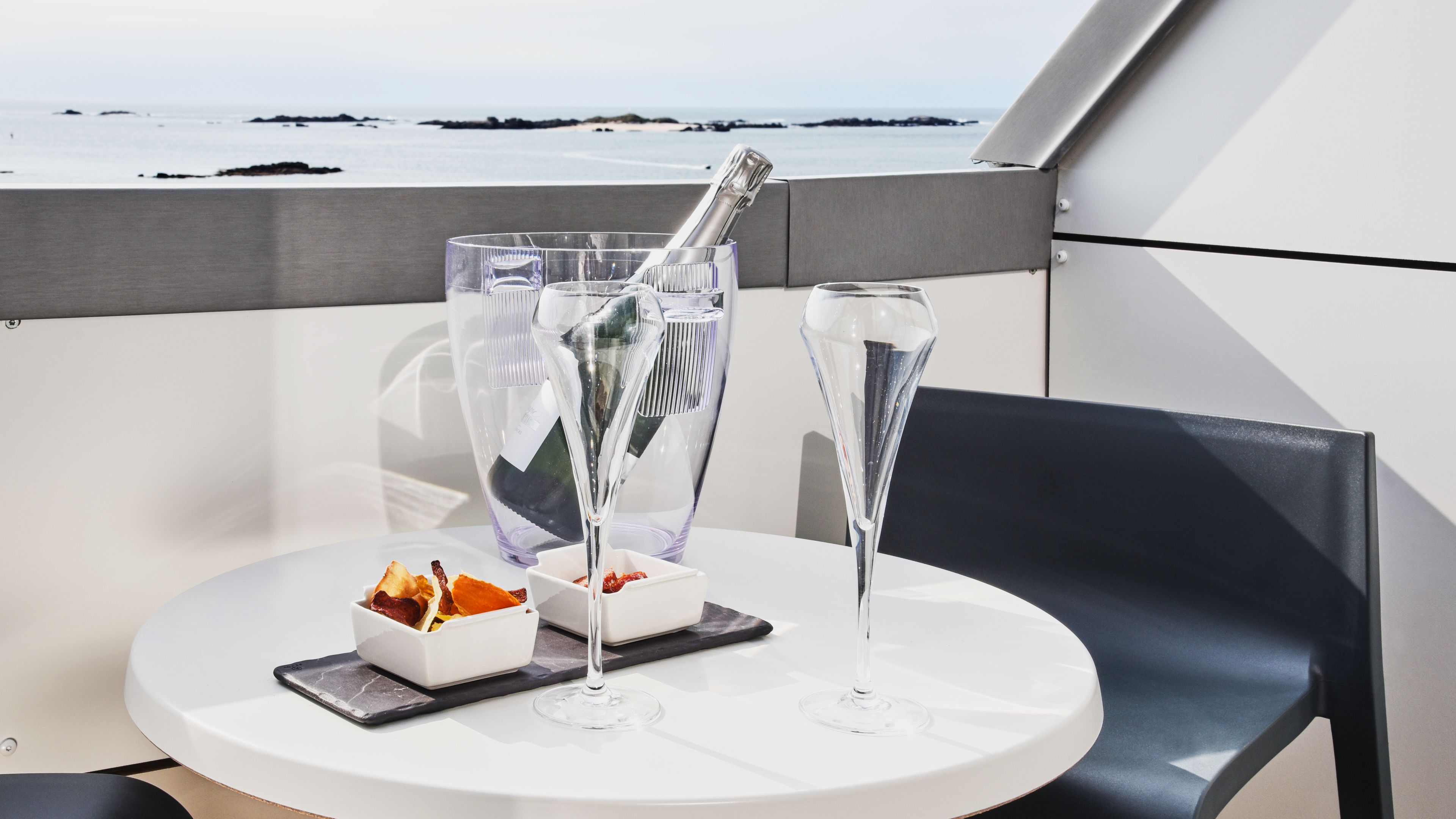 A table with two champagne flutes, a bottle, and a small dish with snacks on a balcony with a view of the sea.