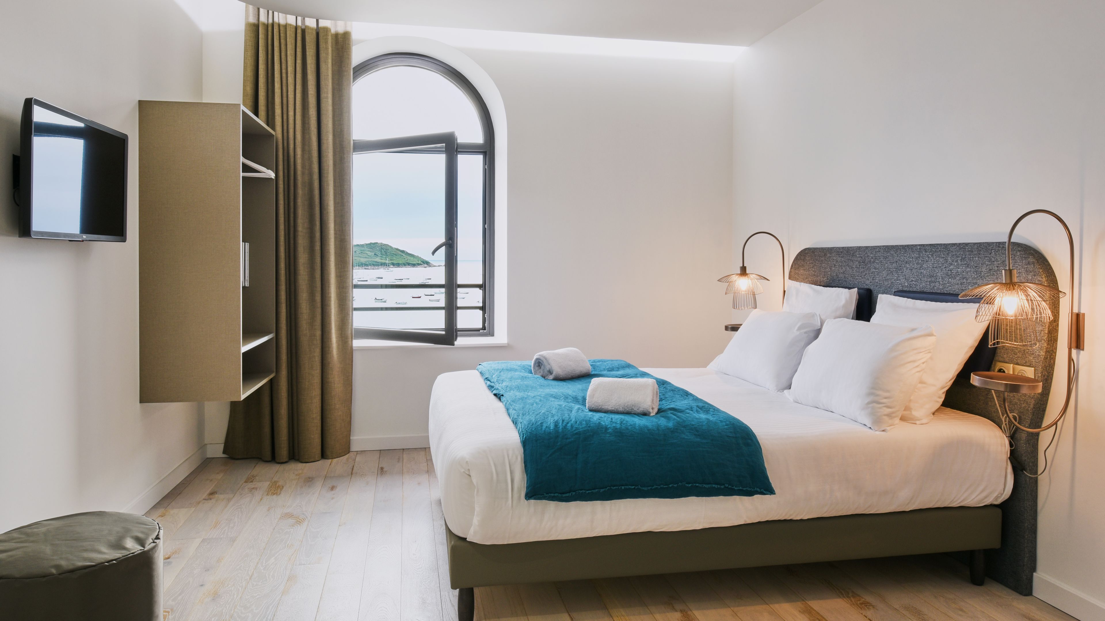 A bedroom with a bed, a TV, a window with curtains, and a view of boats.
