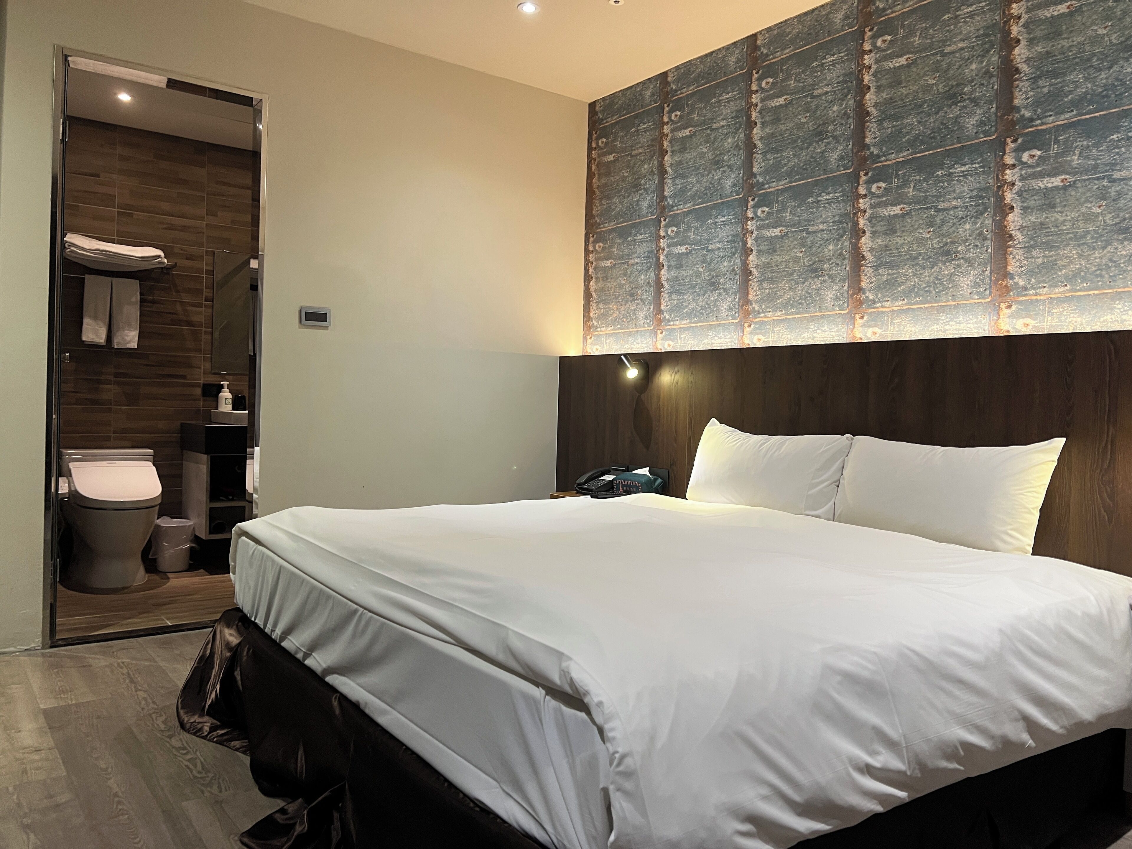 Moderni hotellihuone, jossa on suuri sänky, puinen pääty, sisäänrakennettu penkki, yöpöytä, seinälle asennettu televisio sekä kylpyhuone, jossa on wc ja pesuallas.