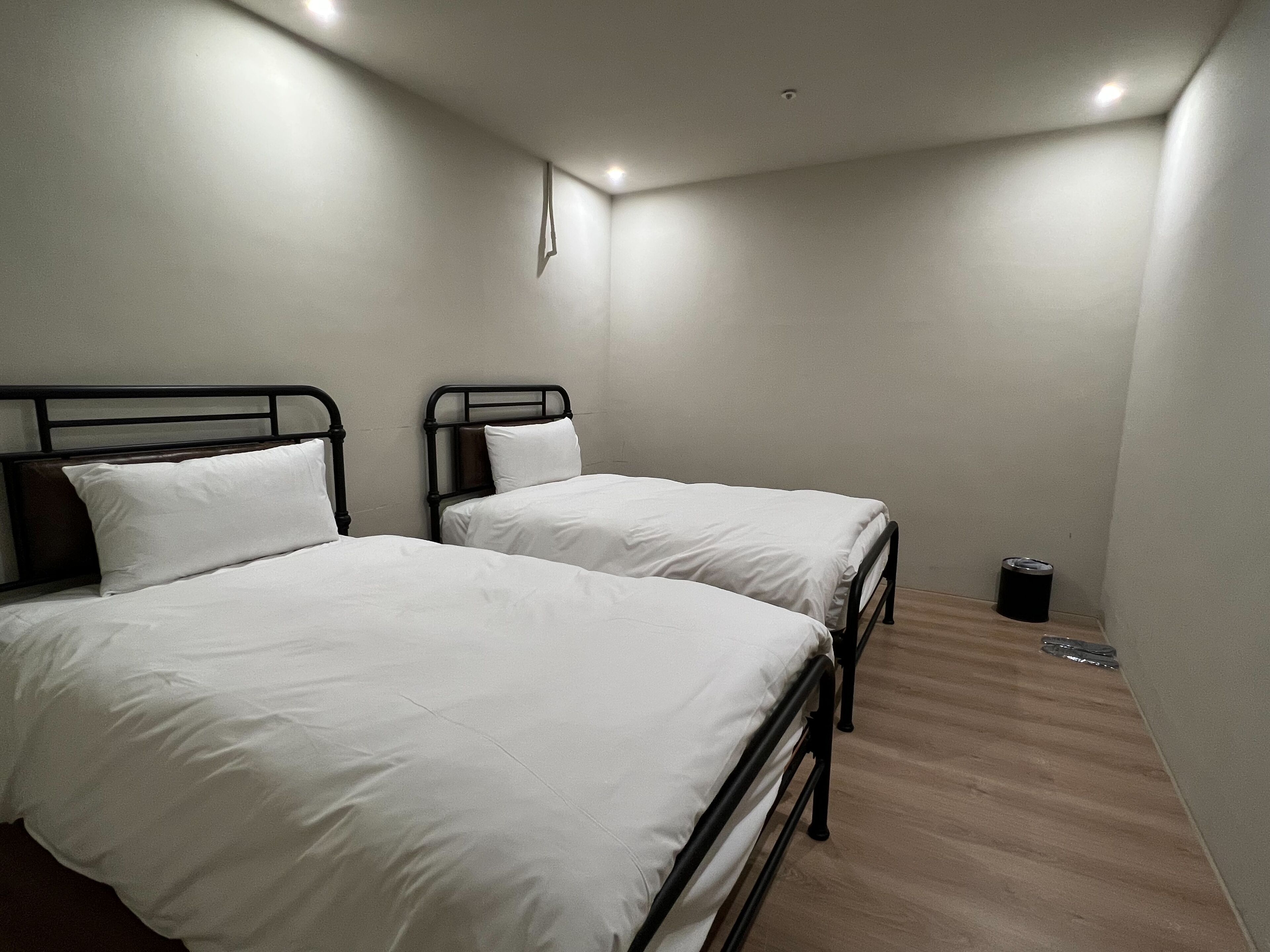 Una habitación con dos camas, cada una con ropa blanca y estructura de metal negro. Hay un cubo de basura pequeño cerca de la pared.