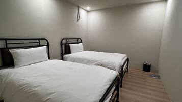 Una habitación con dos camas, cada una con ropa blanca y estructura de metal negro. Hay un cubo de basura pequeño cerca de la pared.