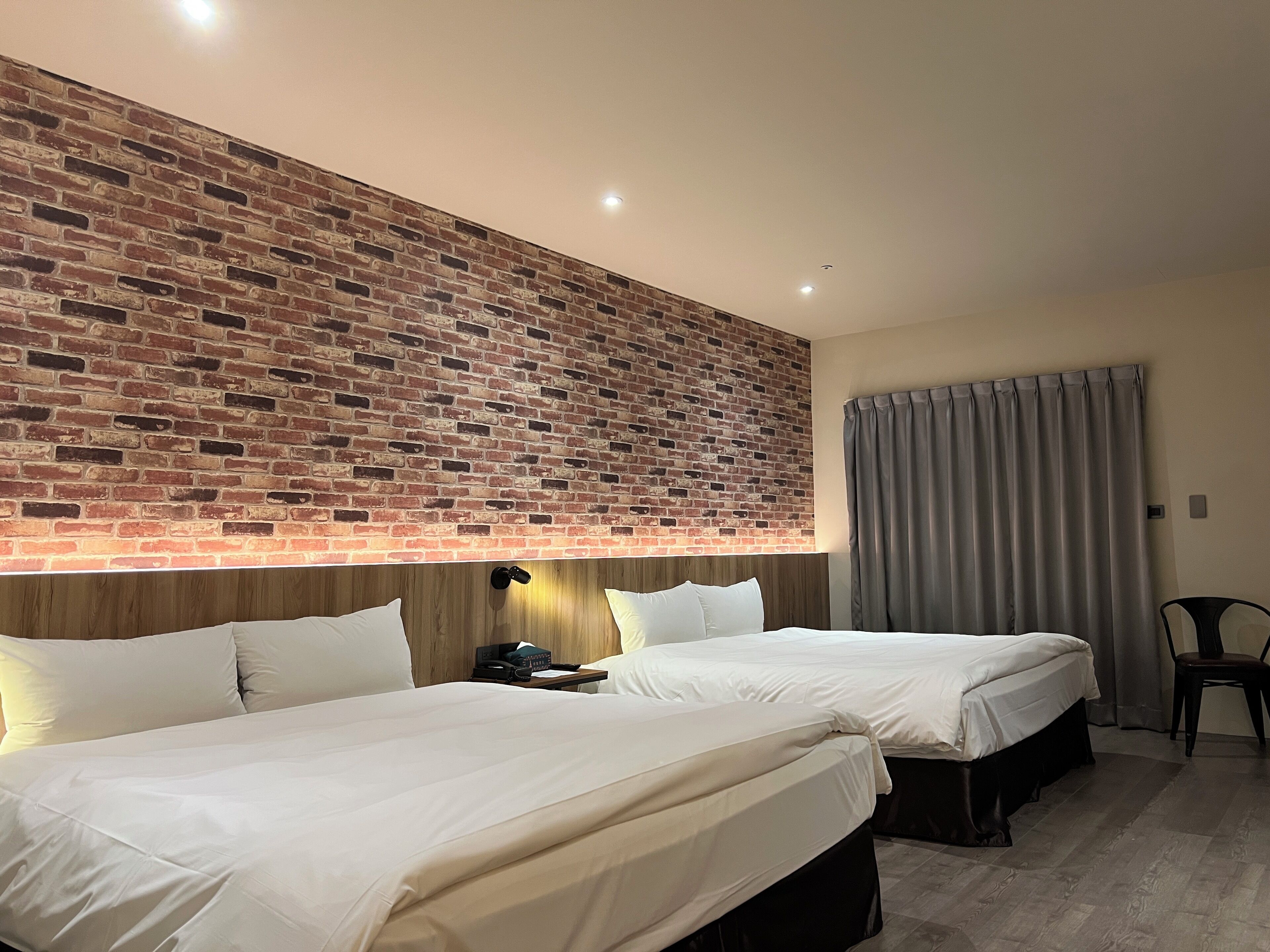 A hotel room with two beds, a brick wall, a wooden headboard, a desk with a lamp, a chair, and a window with curtains.