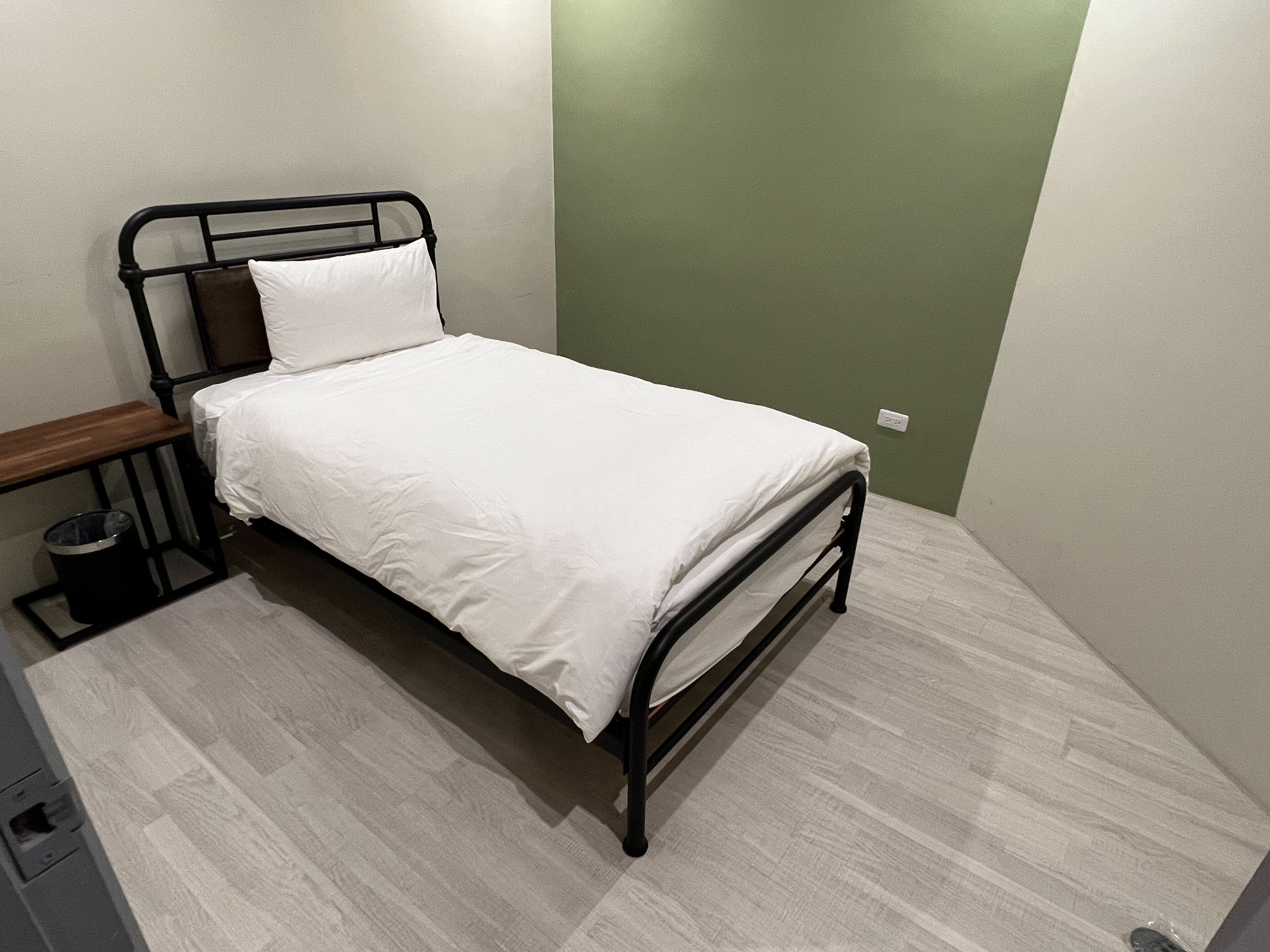 Uma cama de solteiro com estrutura de metal, roupa de cama branca, um banco de madeira e uma lixeira.