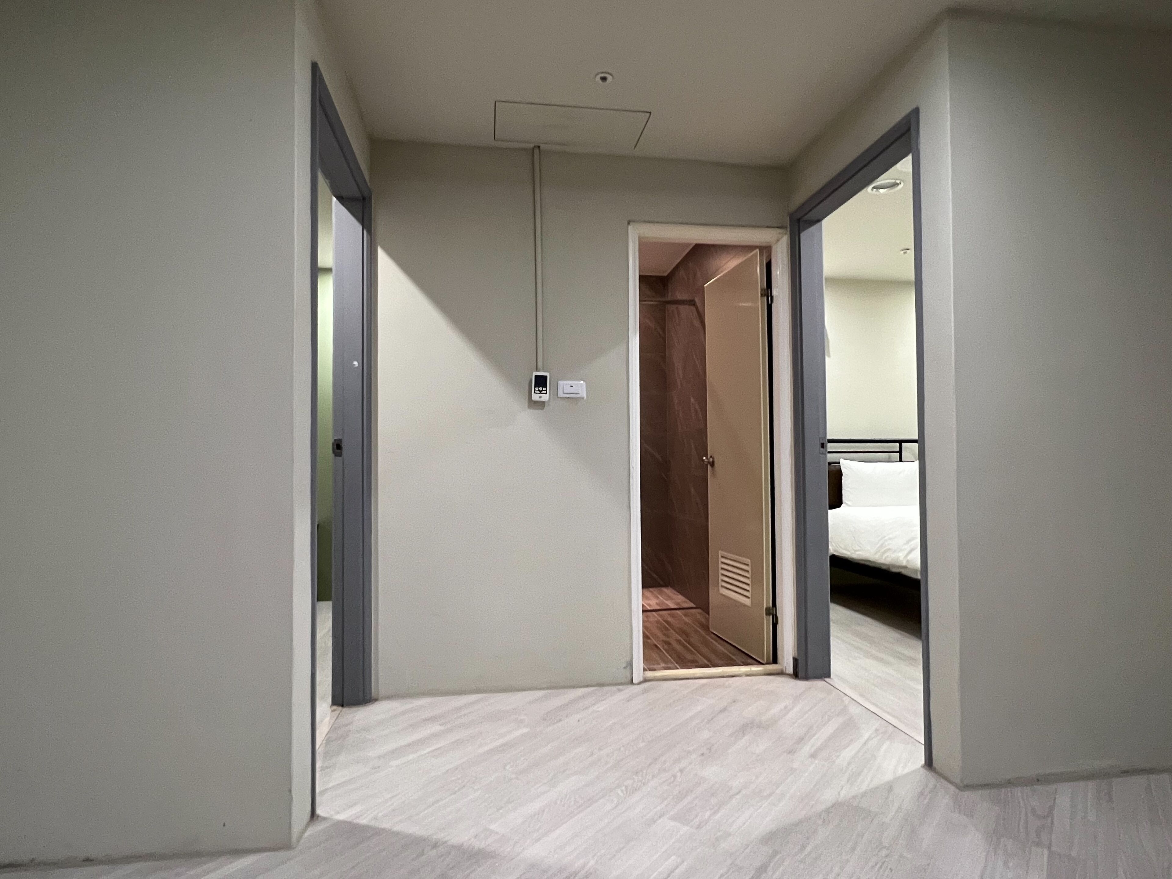 A hotel hallway with a wooden floor, a visible door leading to a room with a bed, and a wall-mounted light fixture.