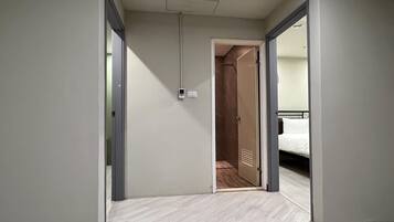 A hotel hallway with a wooden floor, a visible door leading to a room with a bed, and a wall-mounted light fixture.