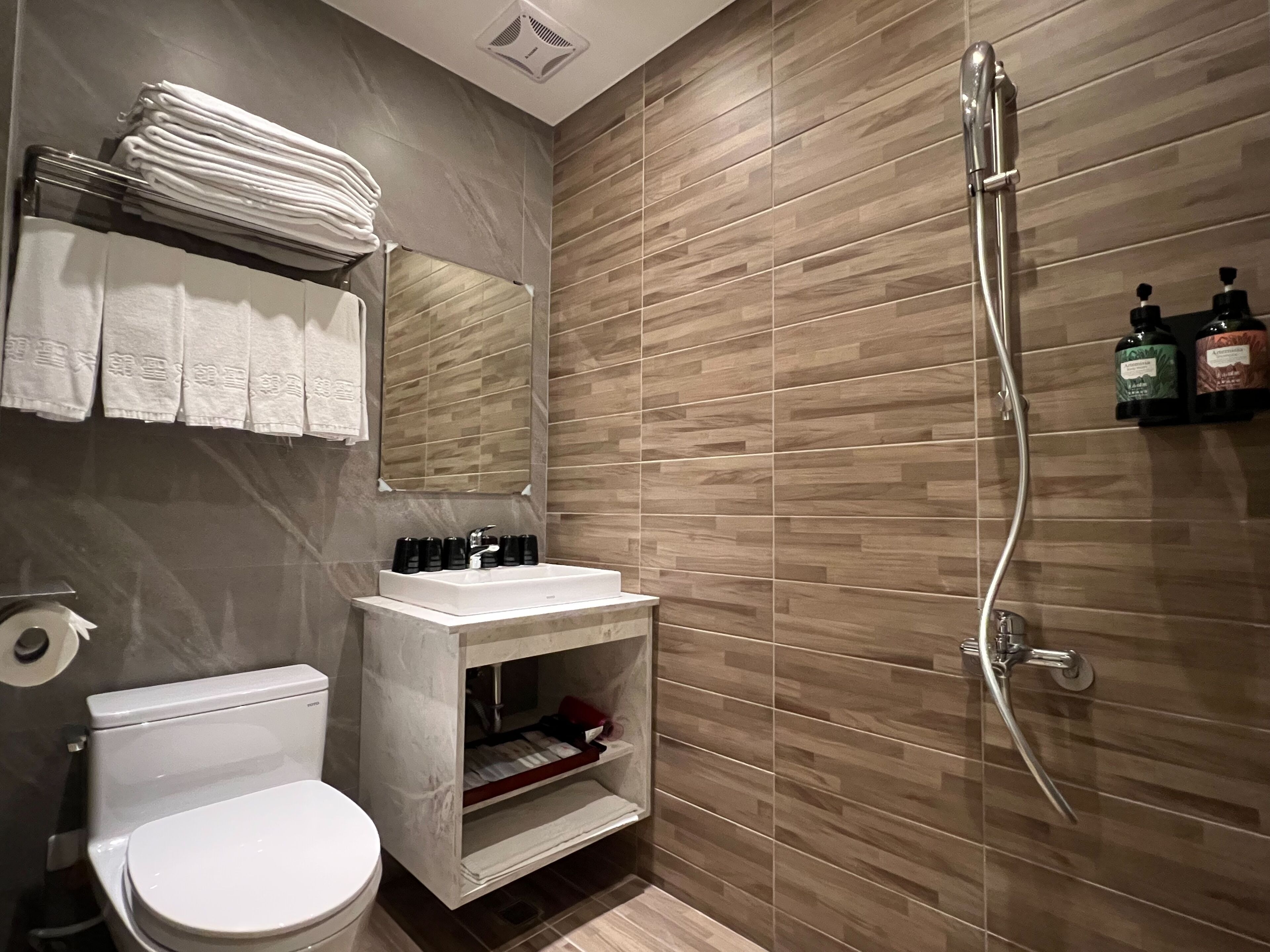 Un baño moderno con inodoro, lavabo y ducha. Hay toallas apiladas en un estante, un espejo sobre el lavabo y artículos de aseo en un estante.