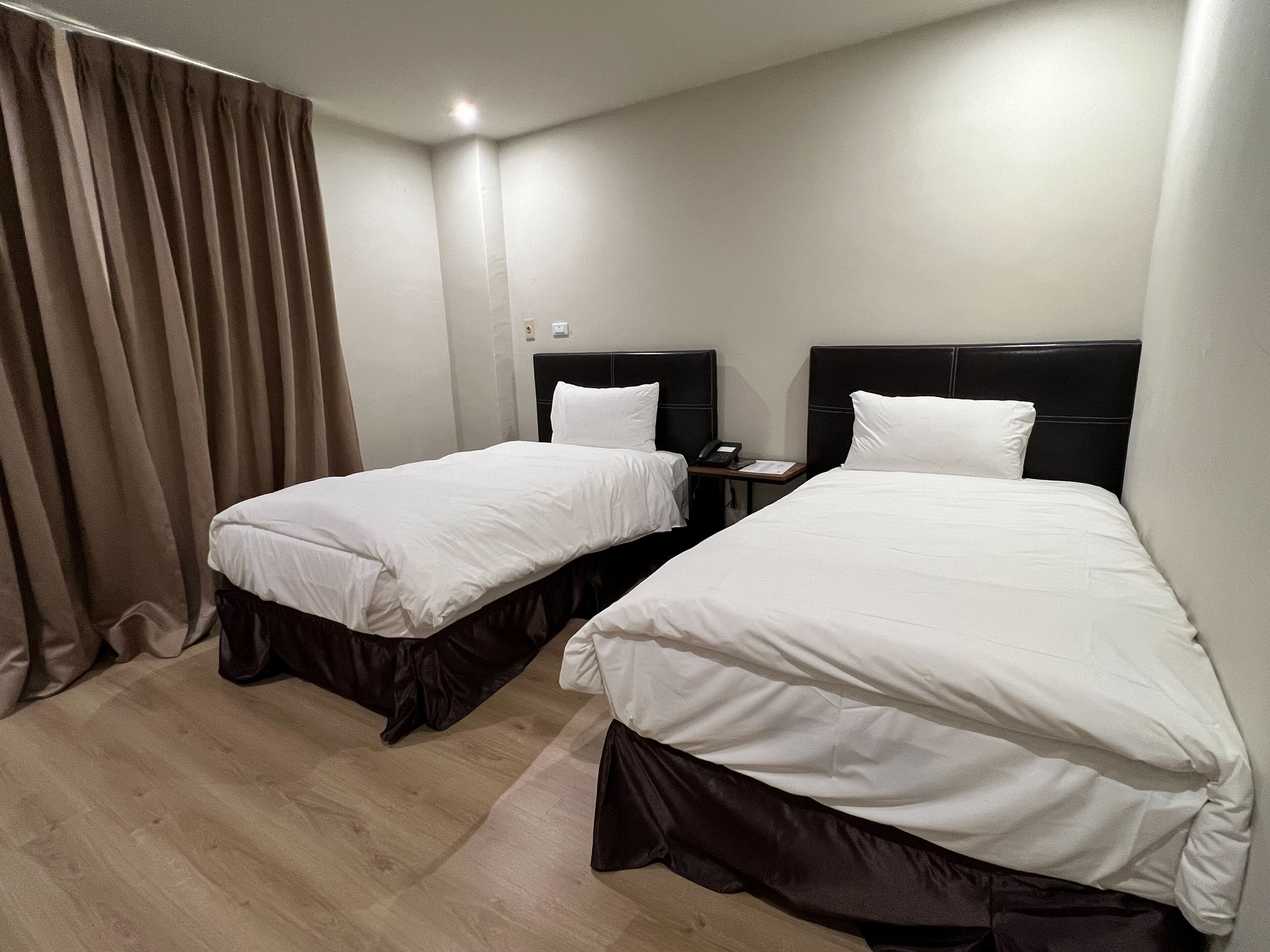 Habitación de hotel con dos camas, cada una con cabecero negro y ropa de cama blanca. Hay una mesita de noche pequeña con un teléfono y una lámpara. El piso es de madera y las cortinas son color beige.