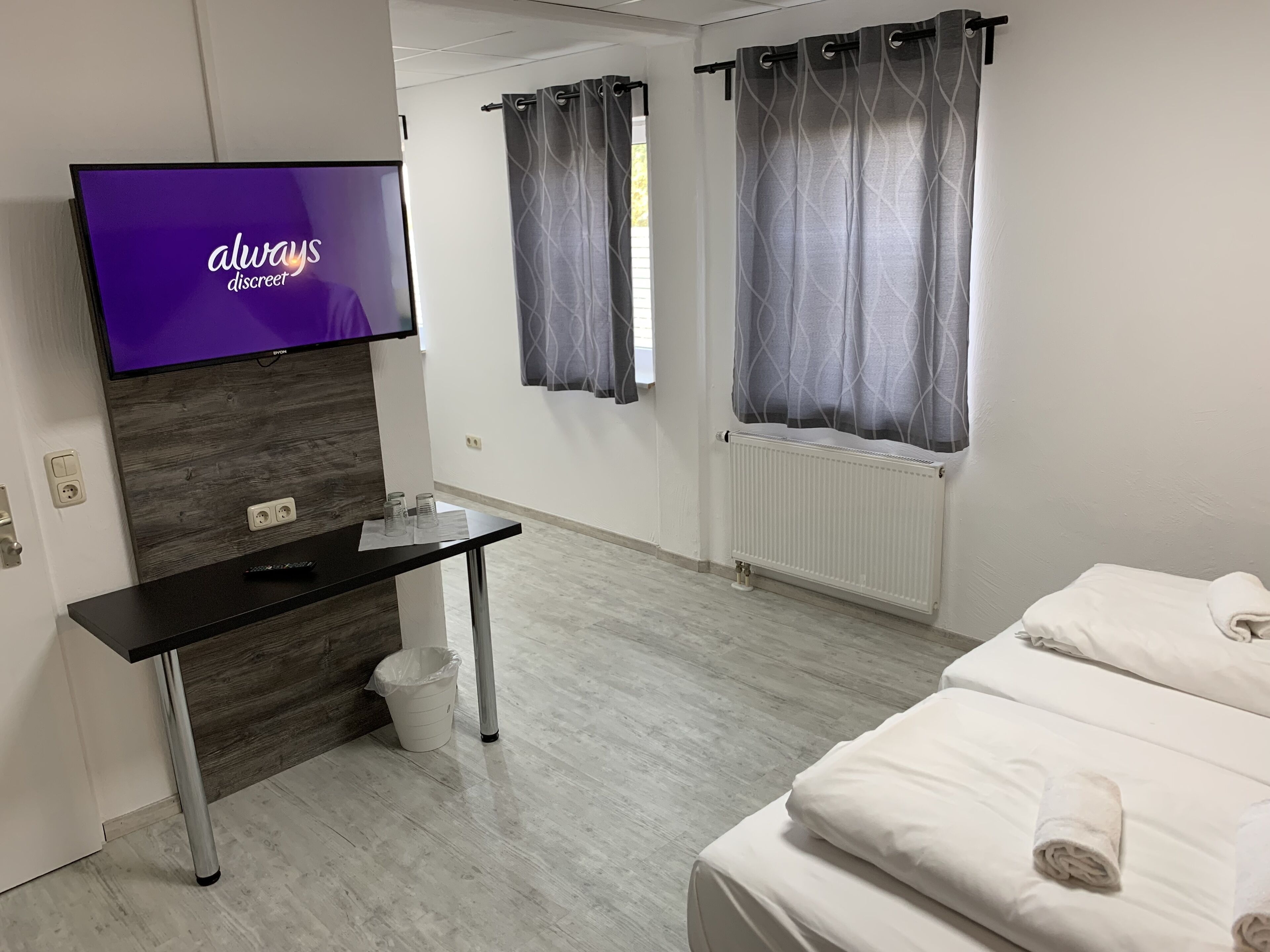 A modern hotel room with a flat-screen TV, a wooden accent wall, a desk with a trash bin, a radiator, and a bed with towels.