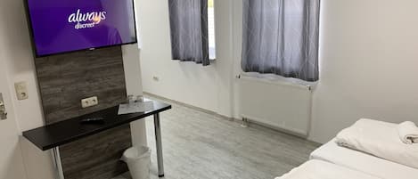 A modern hotel room with a flat-screen TV, a wooden accent wall, a desk with a trash bin, a radiator, and a bed with towels.