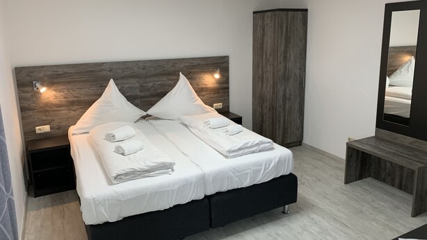 Un dormitorio moderno con cabecera de madera, dos camas con ropa de cama blanca, una mesita de noche, un armario alto y un espejo.