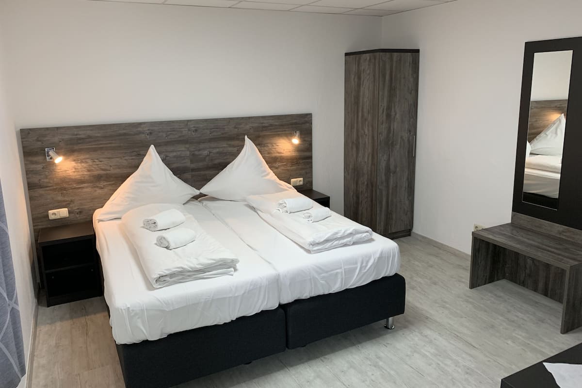 A modern bedroom with a wooden headboard, two beds with white linens, a bedside table, a nightstand, a tall wardrobe, and a mirror.