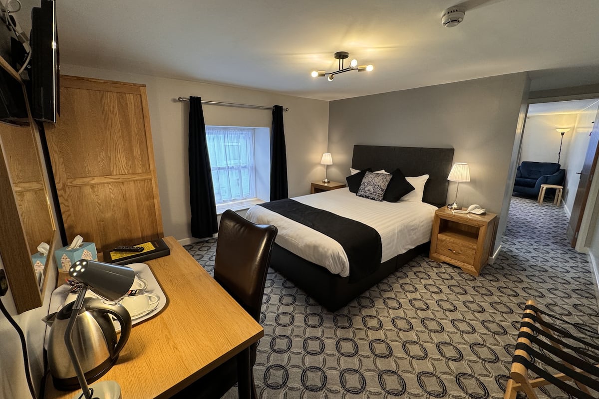 A hotel room with a bed, bedside tables, a desk with a kettle, a chair, a TV, and a patterned carpet.