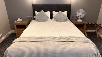 Una cama bien hecha con un cabecero gris, dos mesitas de noche con lámparas, un ventilador y un teléfono a la izquierda.