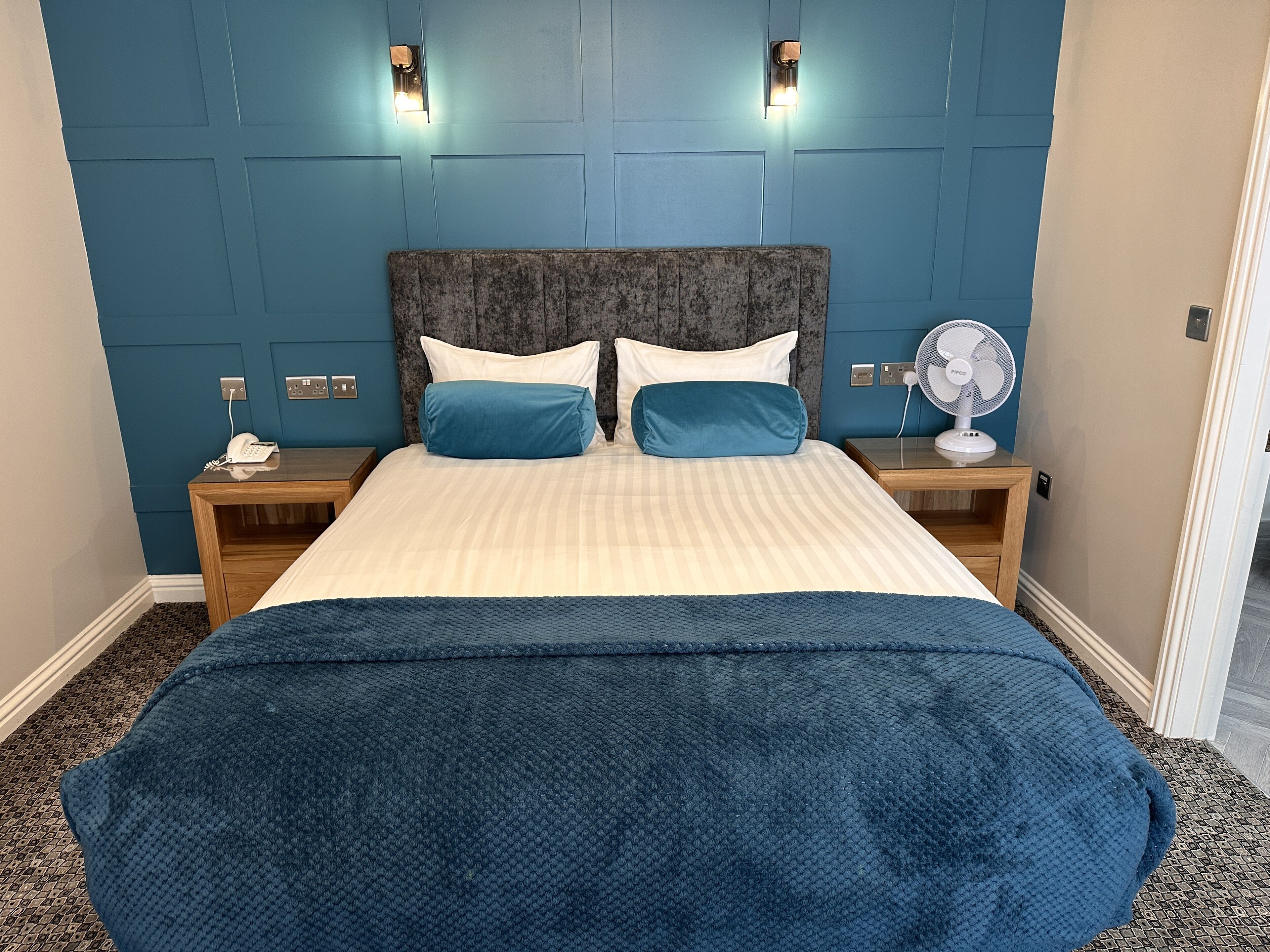 A hotel room with a bed, two bedside tables, a phone, a fan, and wall-mounted lights.
