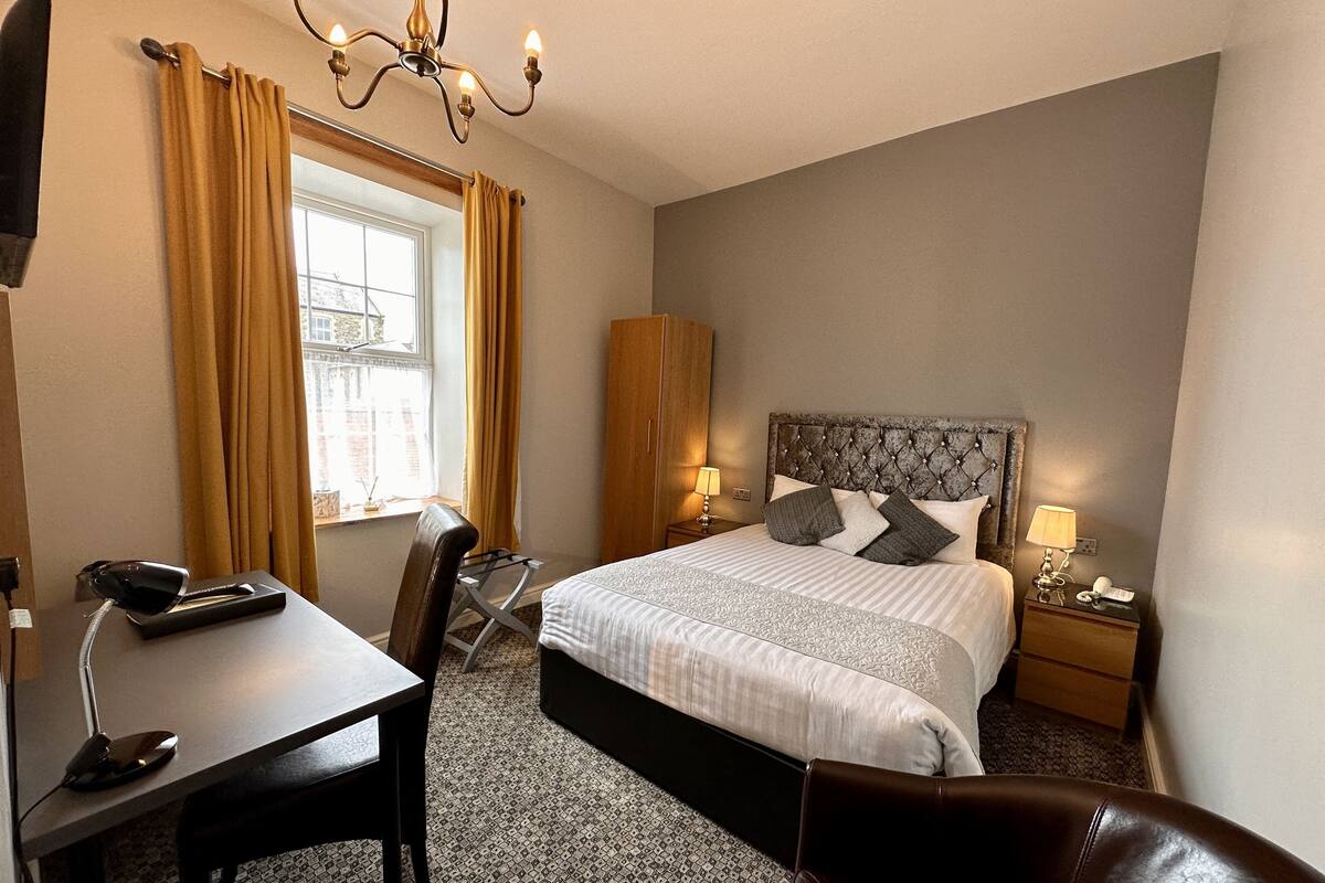 A hotel room with a bed, a desk with a telephone, a chair, a wardrobe, and a chandelier.