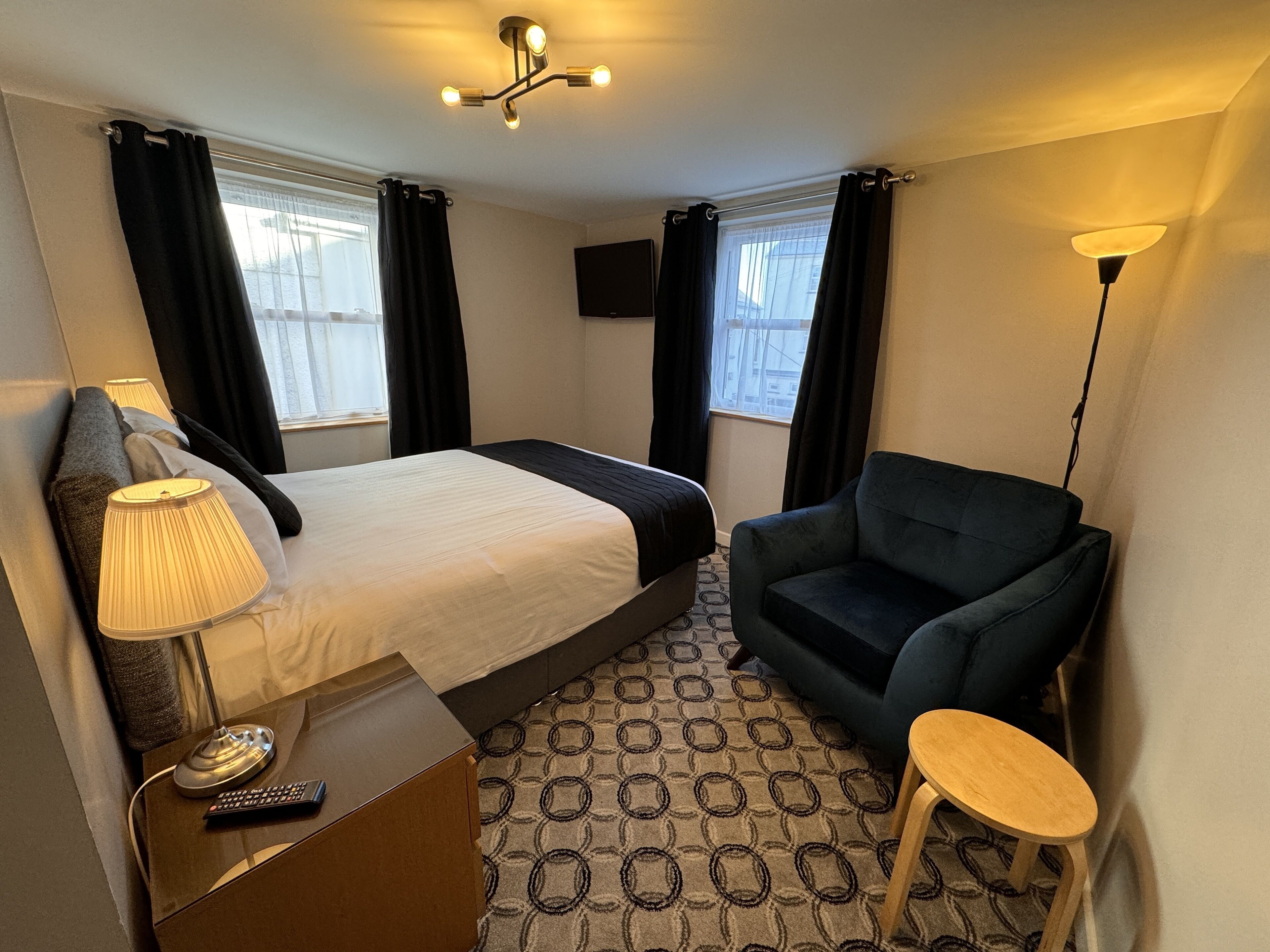 A hotel room with a bed, a desk with a lamp, a chair, a small stool, and a TV on the wall.