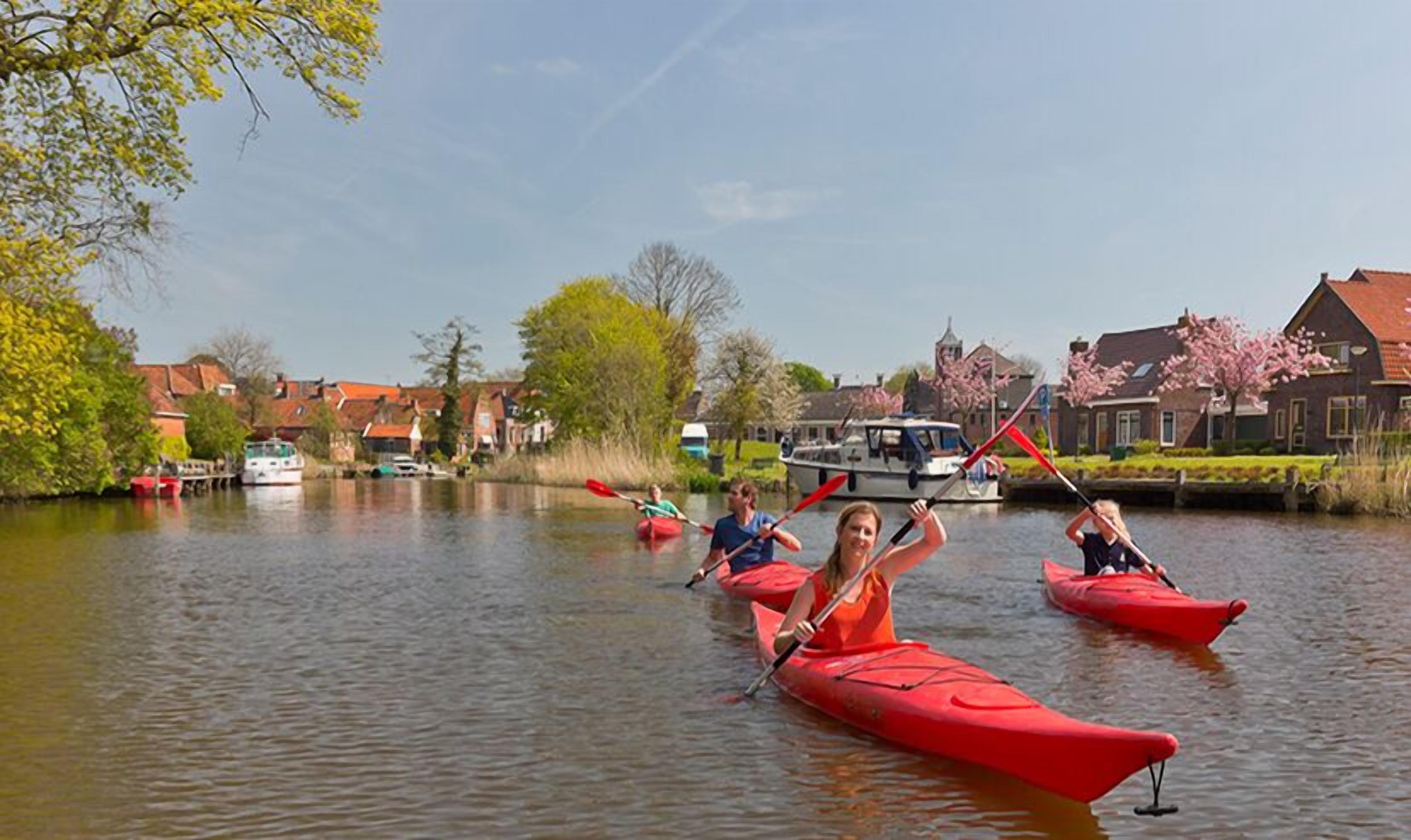 A calm river with people kayaking in red kayaks, surrounded by houses and a boat docked on the riverbank.