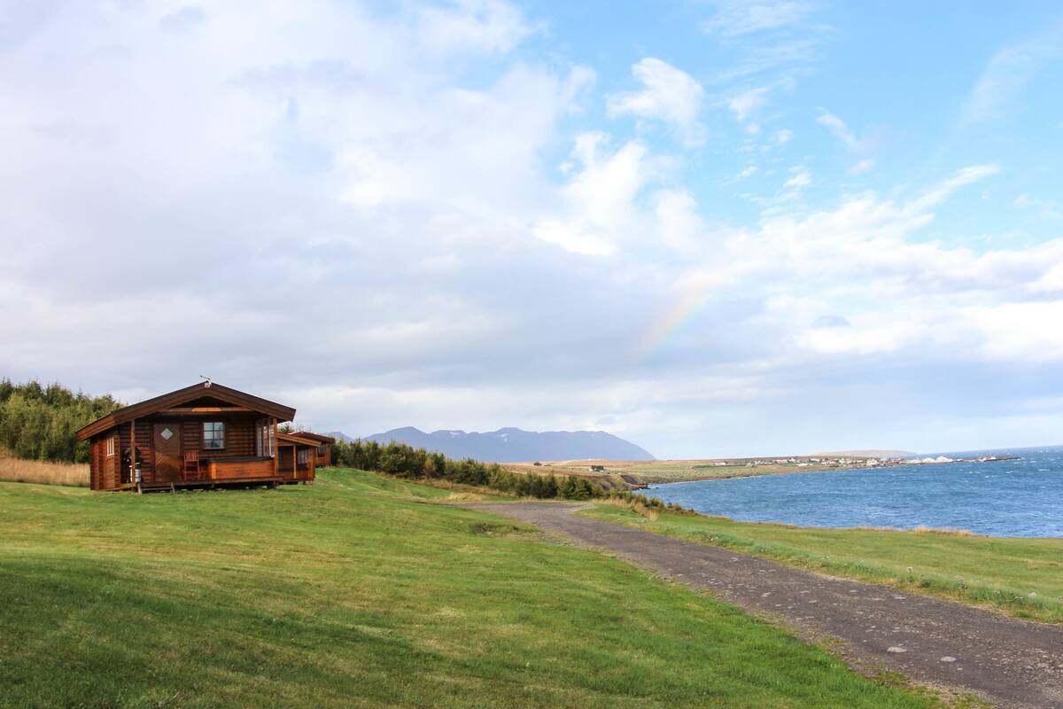 A wooden cabin by a lake with a clear sky and mountains in the background.