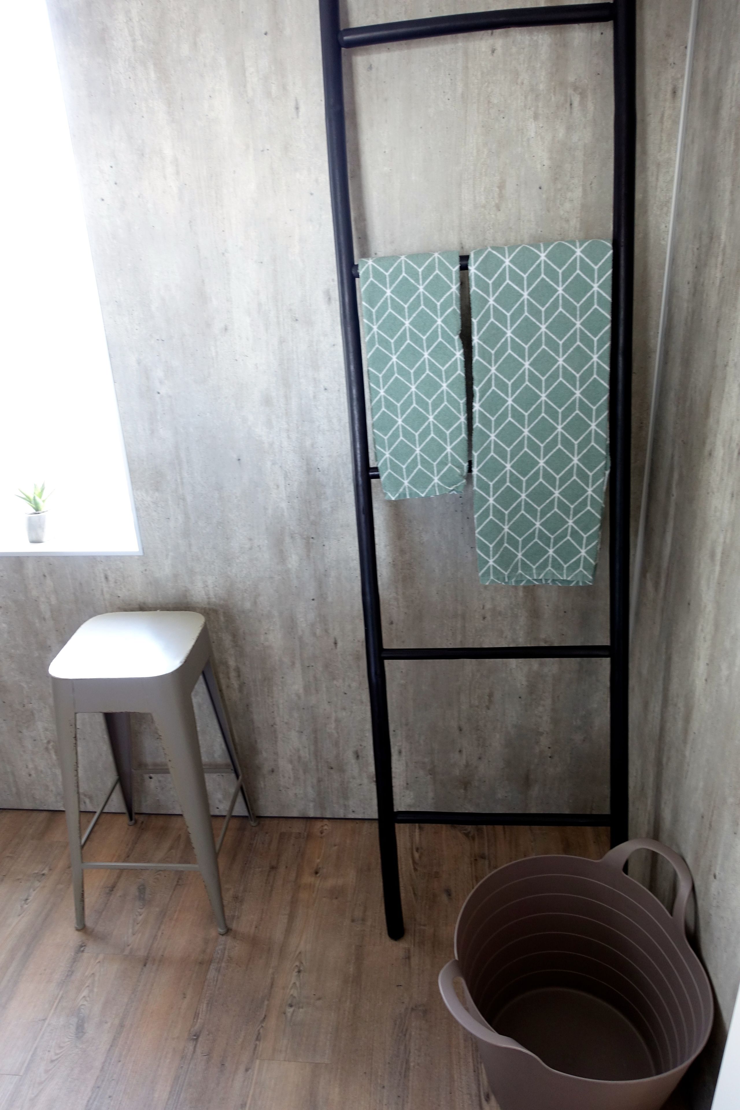 Quarto com parede de concreto, escada de metal, um banquinho e um balde marrom.