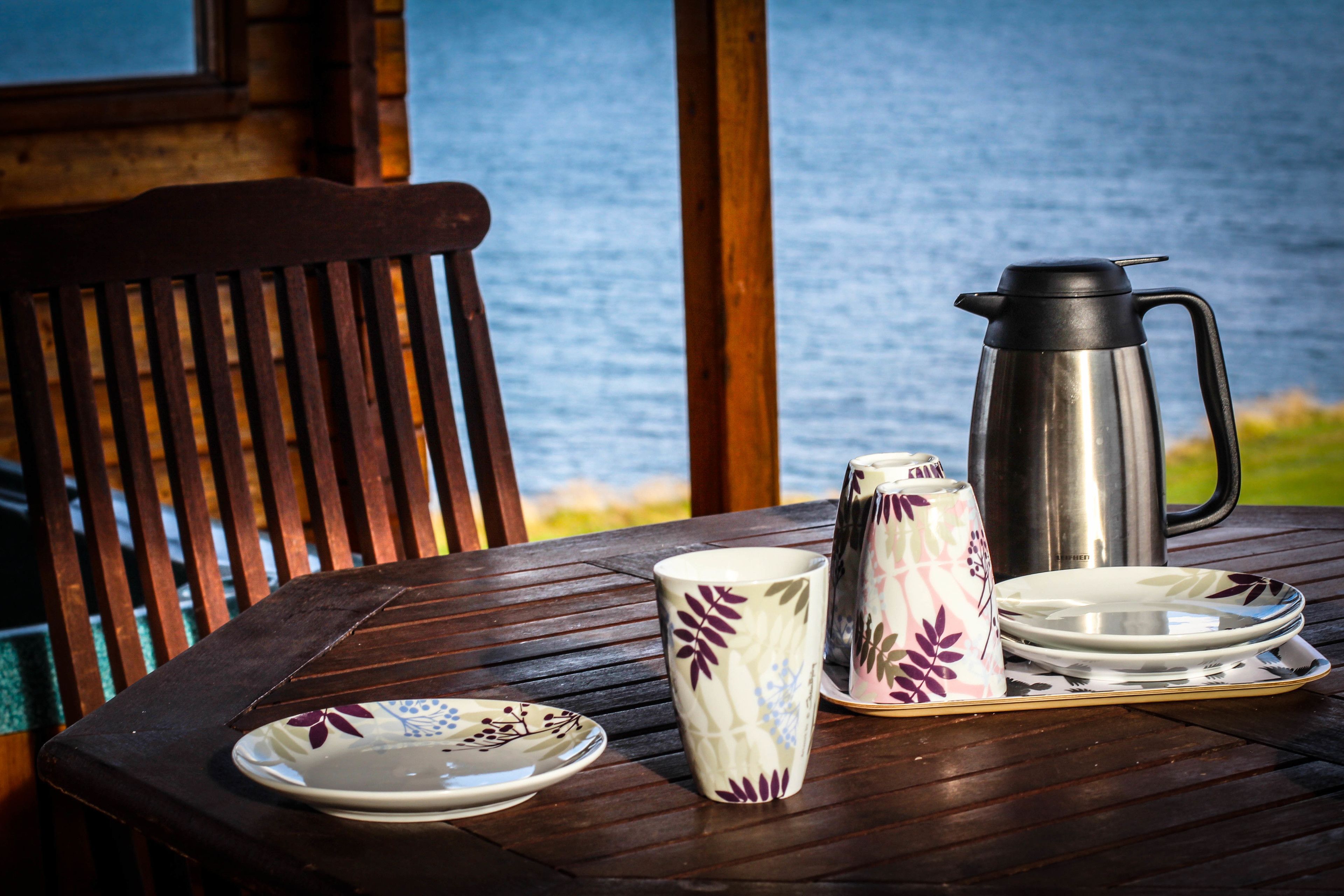 Una mesa de madera con una cafetera metálica, una taza estampada y platos, frente a una vista del mar.
