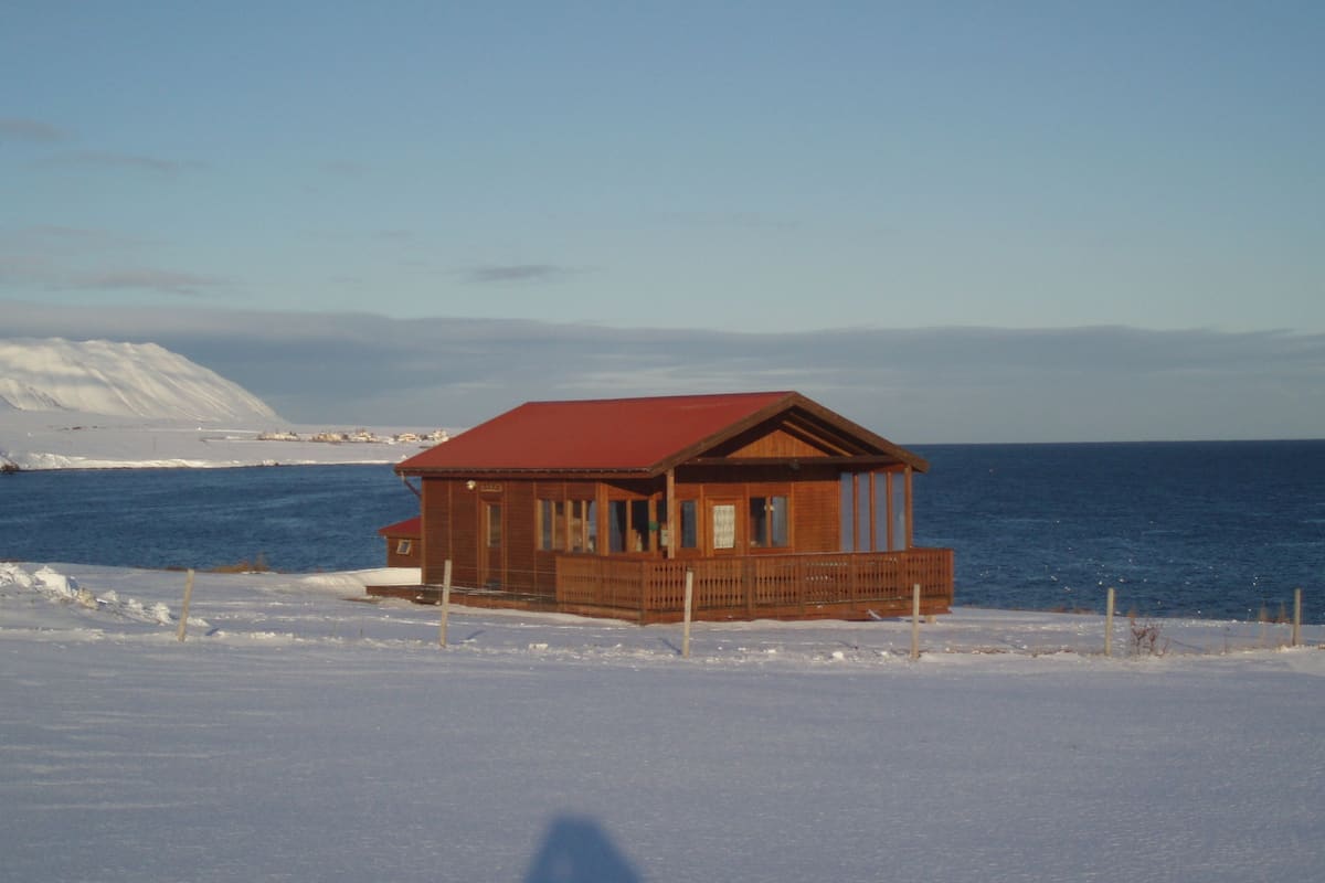 A wooden cabin with a red roof and a balcony, situated on a snow-covered beach with the ocean in the background.