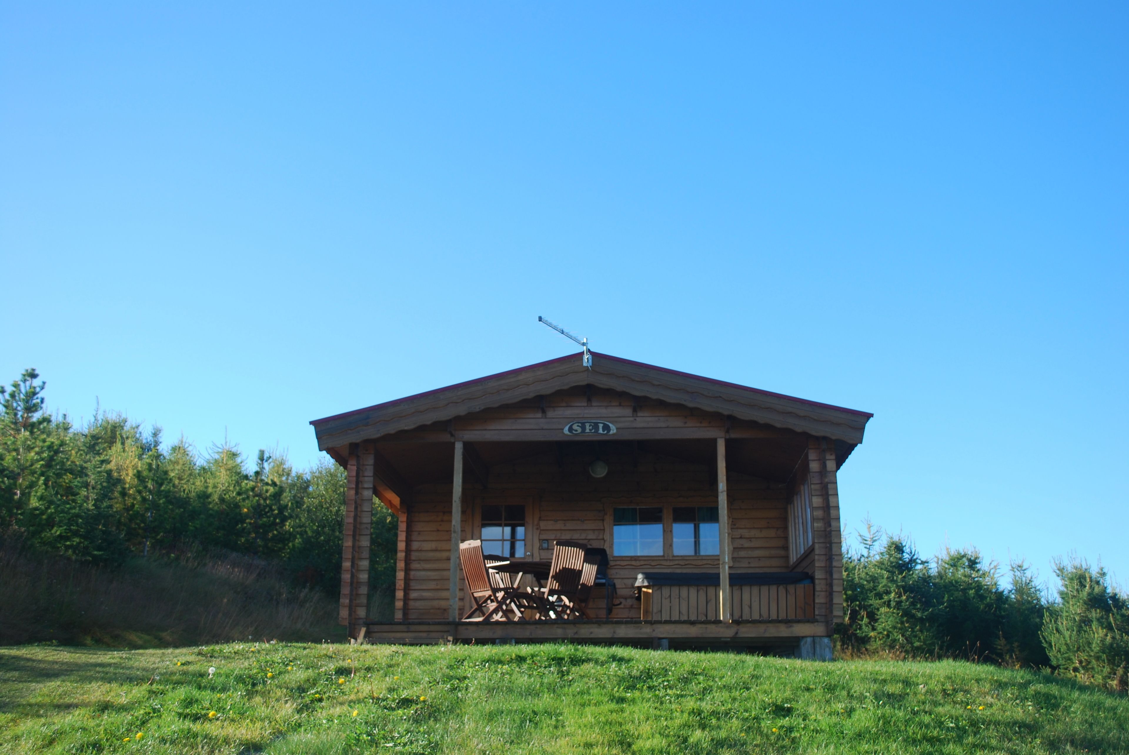 Une cabane en bois avec un porche, portant l’inscription « SEL », entourée d’un champ herbeux et d’arbres.
