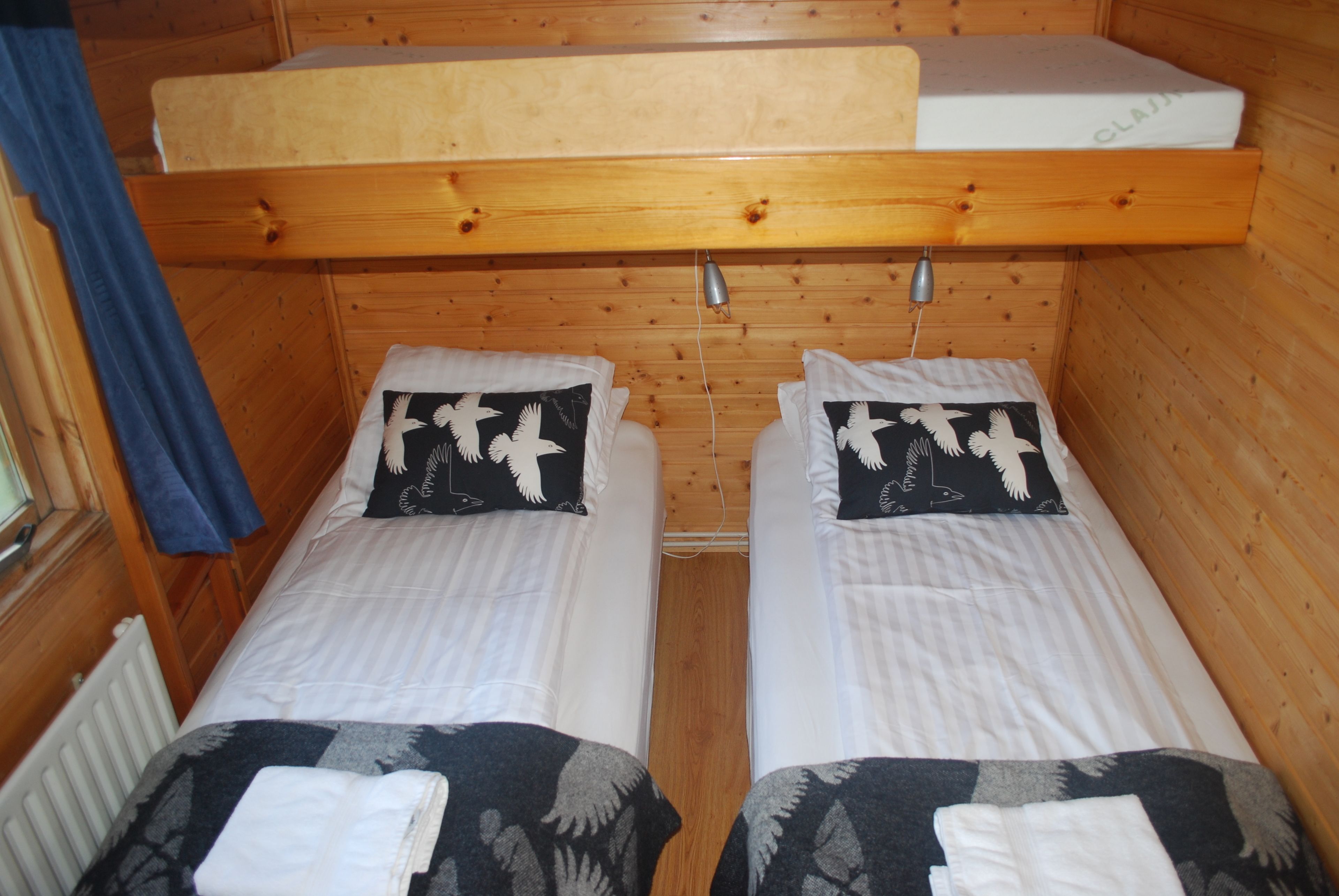 A cabin-style room with two single beds, each with a pillow featuring a white bird design. There is a wooden bunk bed above the beds, and a window with a blue curtain on the left.