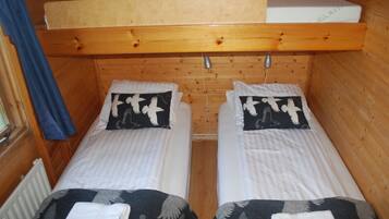 A cabin-style room with two single beds, each with a pillow featuring a white bird design. There is a wooden bunk bed above the beds, and a window with a blue curtain on the left.