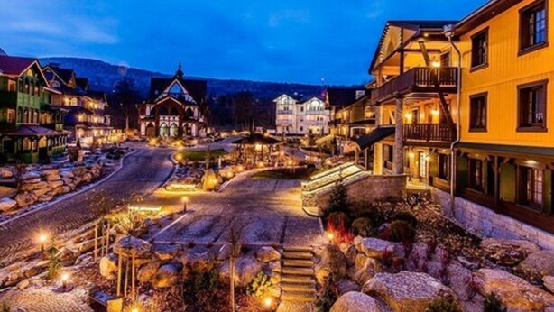 Une place de ville bordée de bâtiments, une rue pavée et une chaîne de montagnes en arrière-plan.