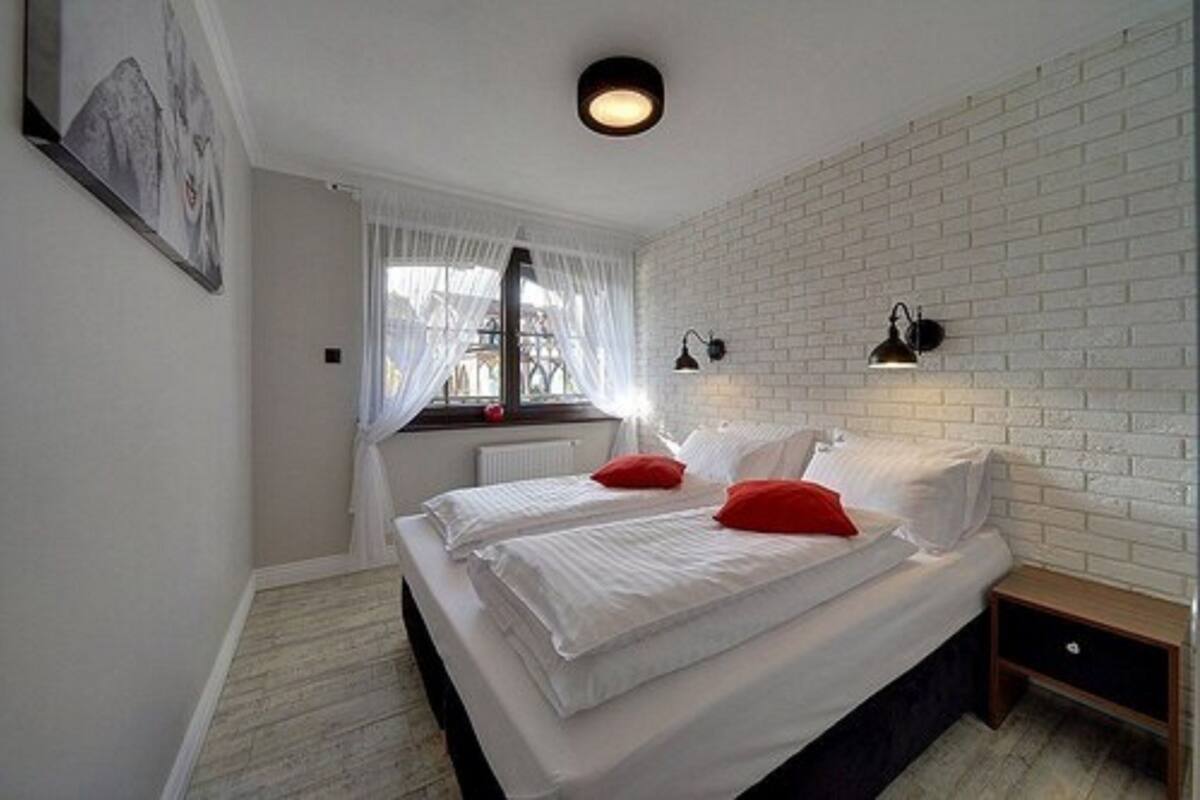 A bedroom with a white brick wall, a bed with white bedding and red pillows, a window with sheer curtains, and two wall-mounted lamps.