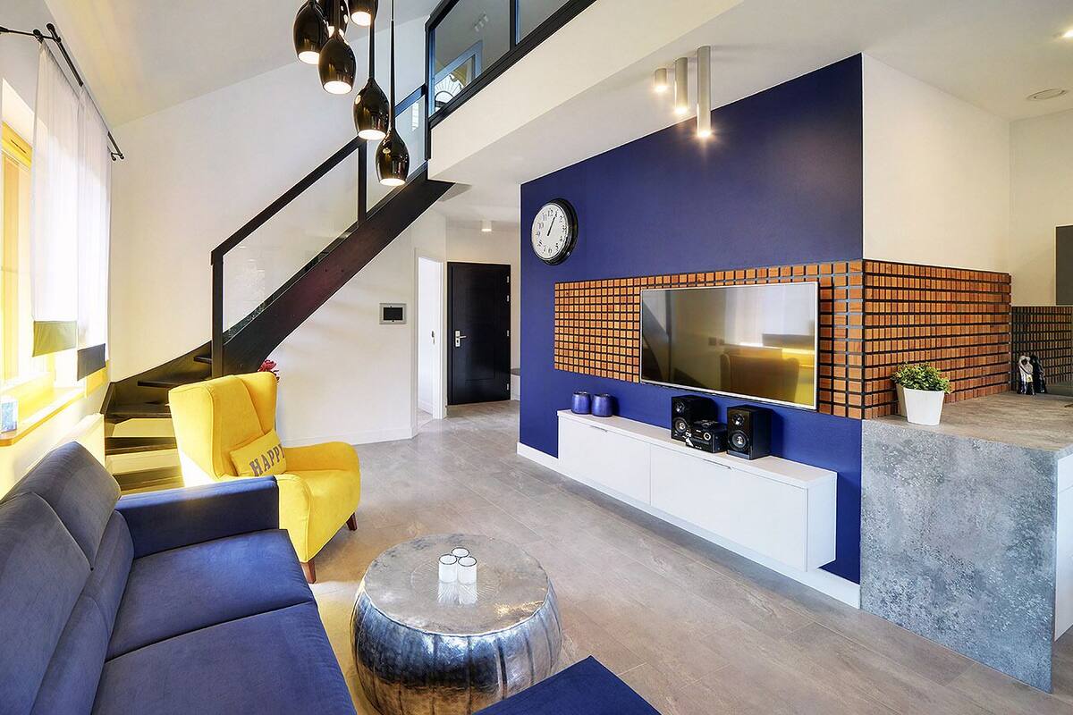 A modern living room with a staircase, a sofa, a coffee table, and a kitchen area with a TV and speakers.