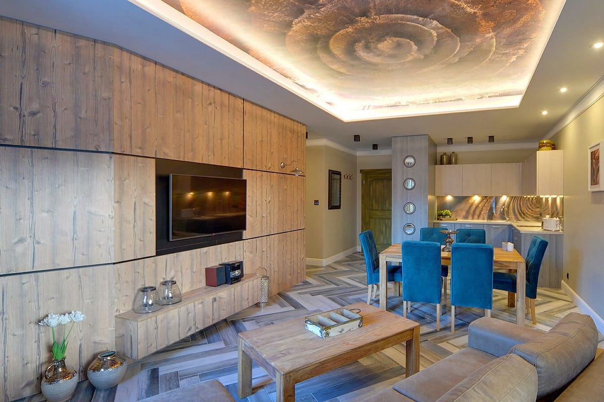 A modern living room with a wooden cabinet, a TV, a dining area with blue chairs, and a kitchen in the background.