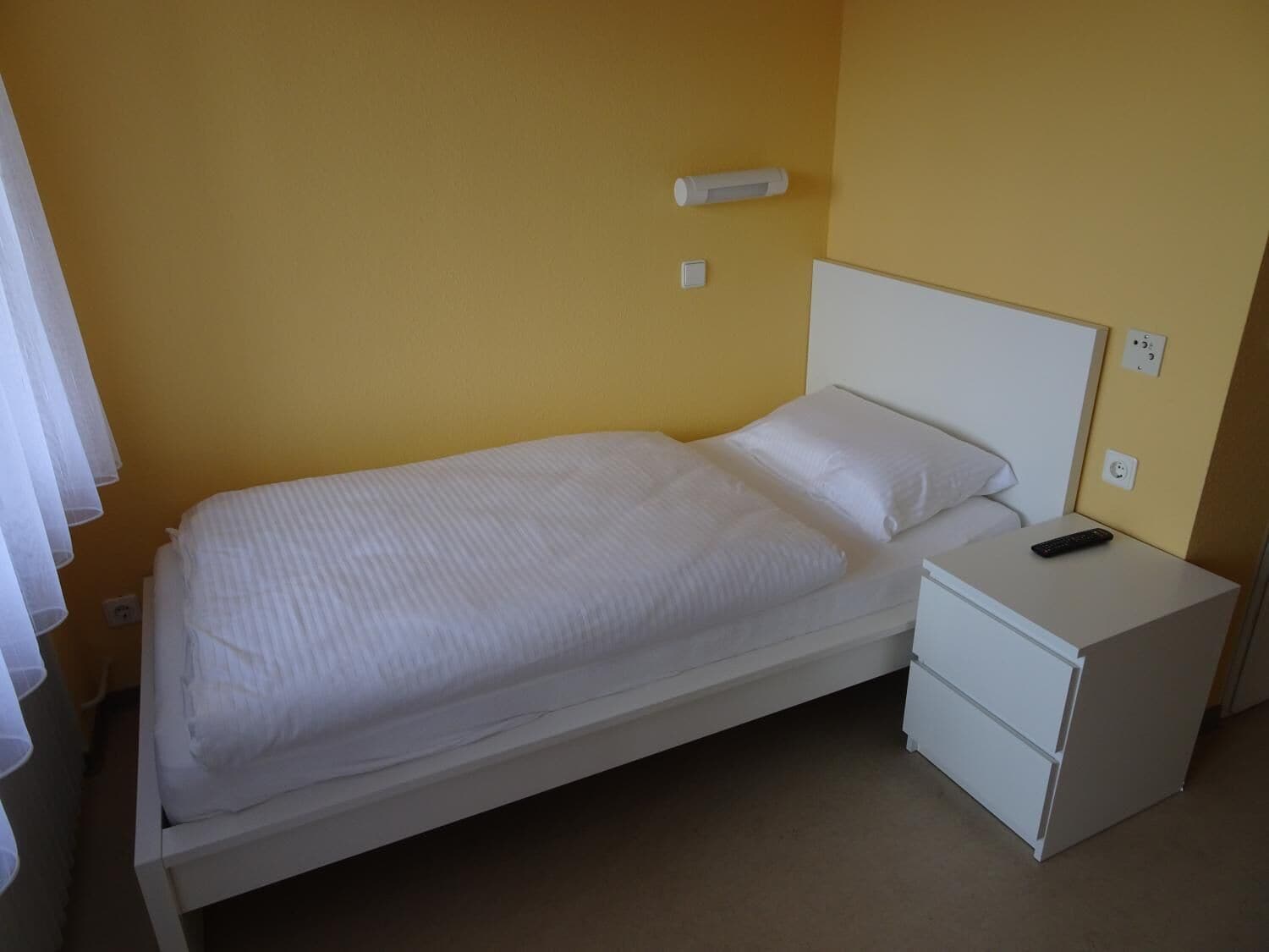 A single-bed room with a white bed, a nightstand, and a remote control on it.
