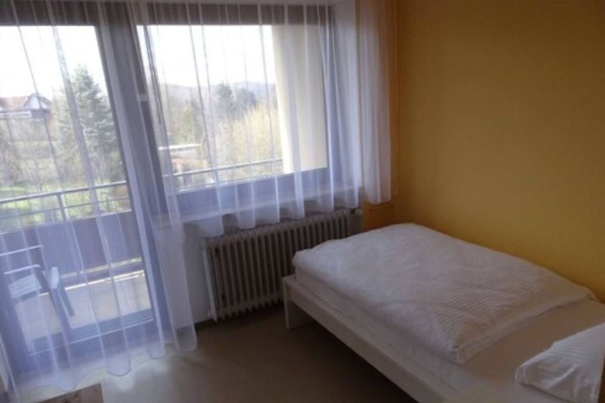 Een kleine, eenvoudige kamer met een bed, een radiator en een raam met dunne gordijnen.