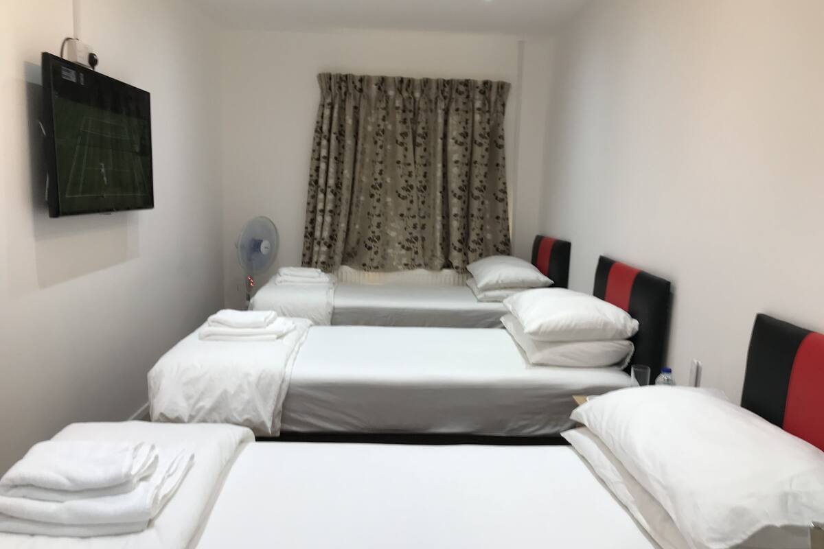 A hotel room with two single beds, a large bed, a flat-screen TV, a fan, and floral curtains.