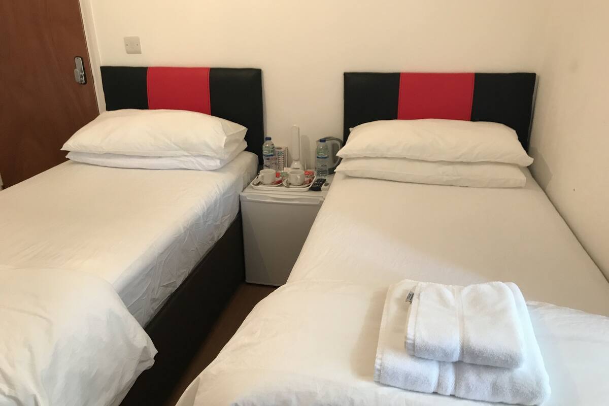 Two single beds with white linens and red and black headboards, a small bedside table with a kettle, cups, and a bottle, and a folded towel on the right bed.