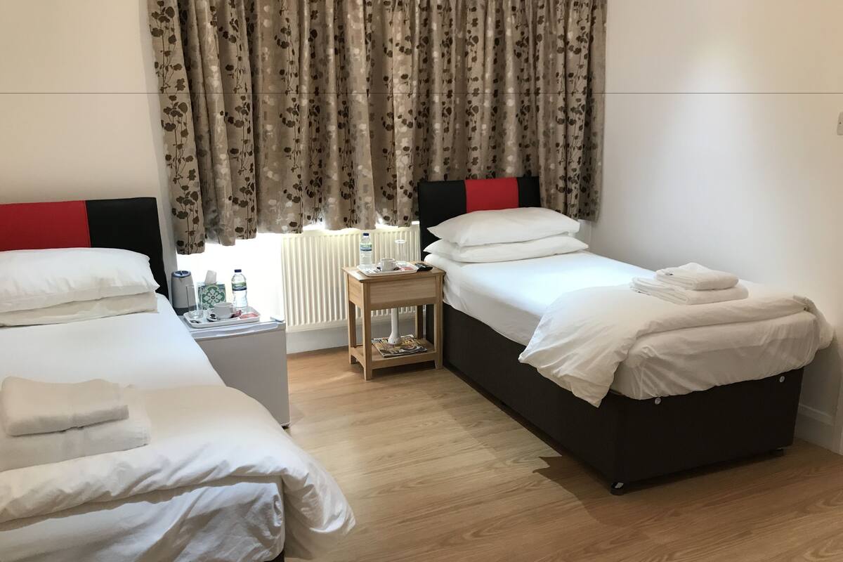A hotel room with two single beds, a small wooden nightstand, a radiator, and floral curtains.
