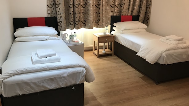 A hotel room with two single beds, a wooden nightstand with a lamp, a small table with a water bottle and a cup, and floral curtains. - Oxford Bed and Breakfast (Kidlington)