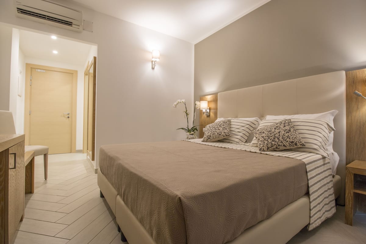 A bedroom with a large bed, bedside tables, a nightstand, a potted plant, and a door leading to another room.