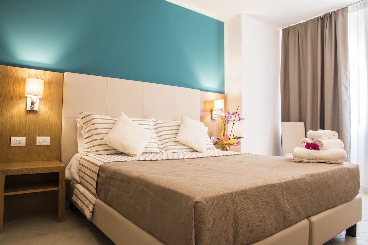 A hotel room with a large bed, a nightstand, a lamp, a vase with flowers, and a chair with towels.