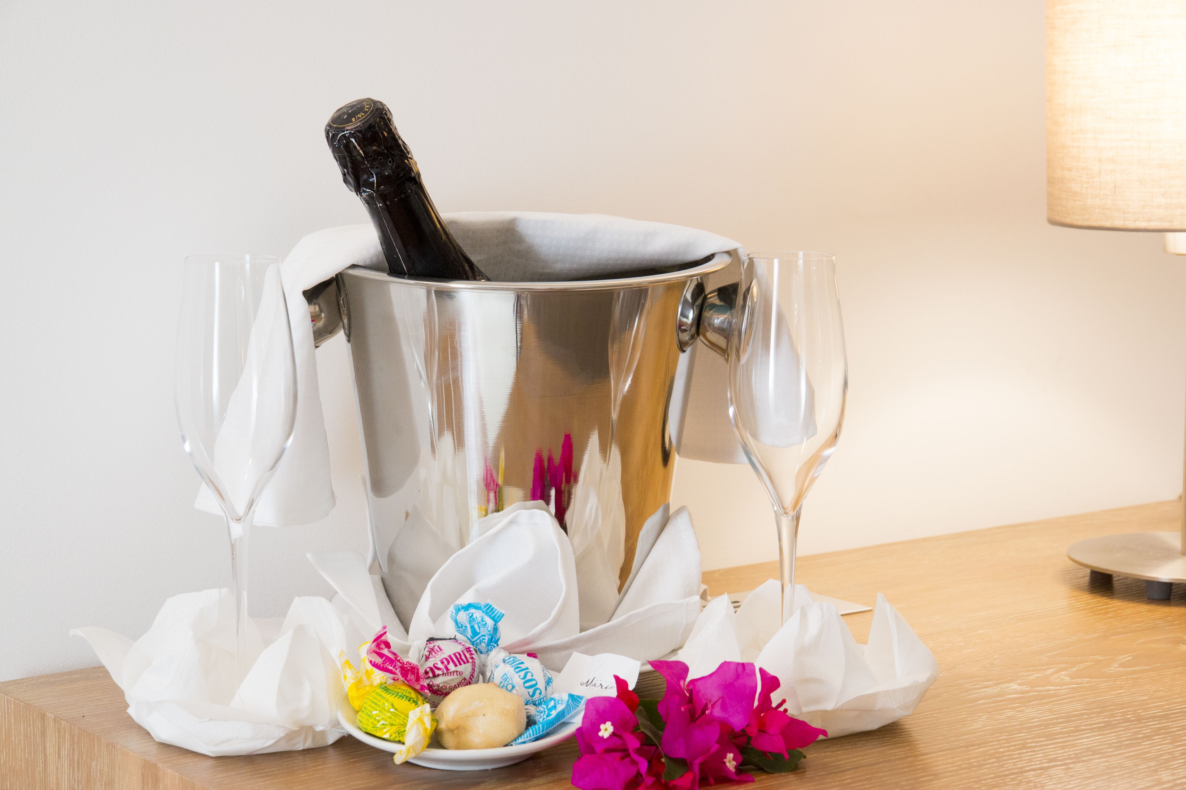 A champagne bottle in a metallic bucket, surrounded by empty champagne flutes, a plate with chocolates, and a flower on a wooden table.