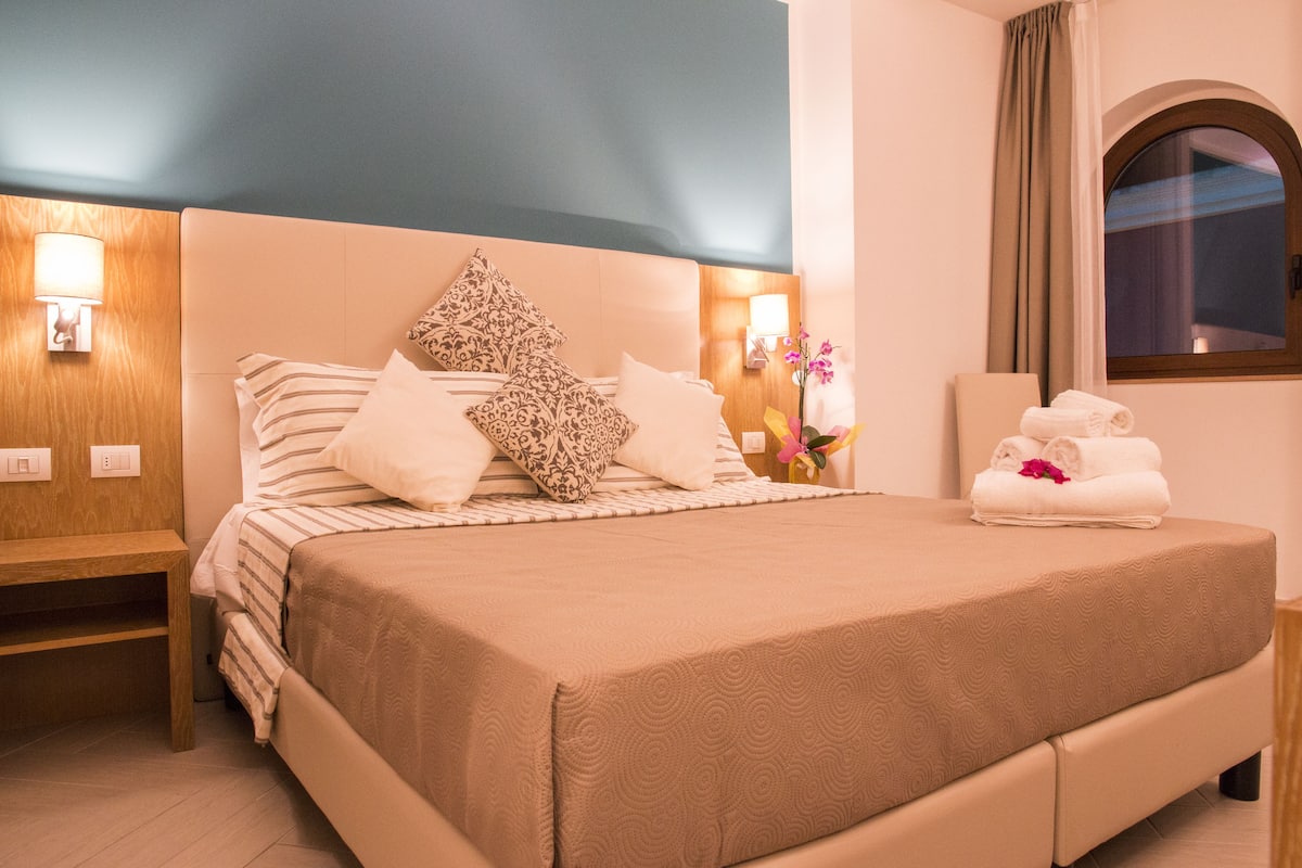 A neatly made bed with patterned pillows, a bedside table with a lamp, and a window with curtains.