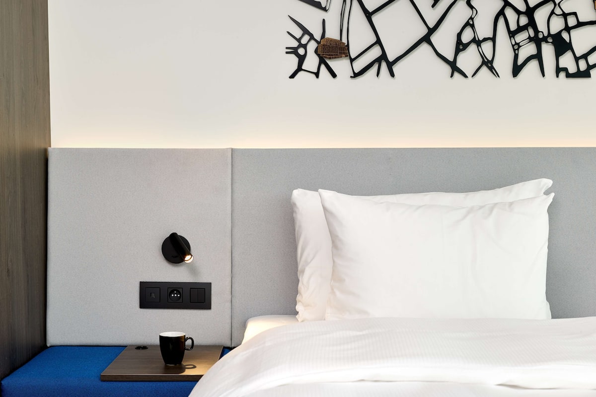 A modern hotel room with a bed, a nightstand, a coffee cup, and a wall-mounted light fixture.