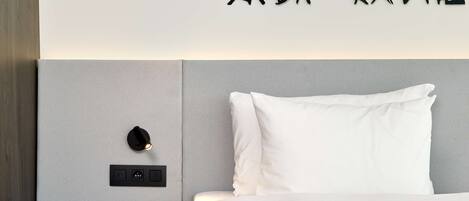A modern hotel room with a bed, a nightstand, a coffee cup, and a wall-mounted light fixture.